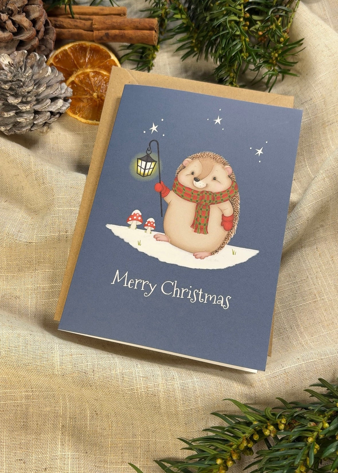 Christmas card with a hedgehog illustration on a blue background, surrounded by festive decorations.